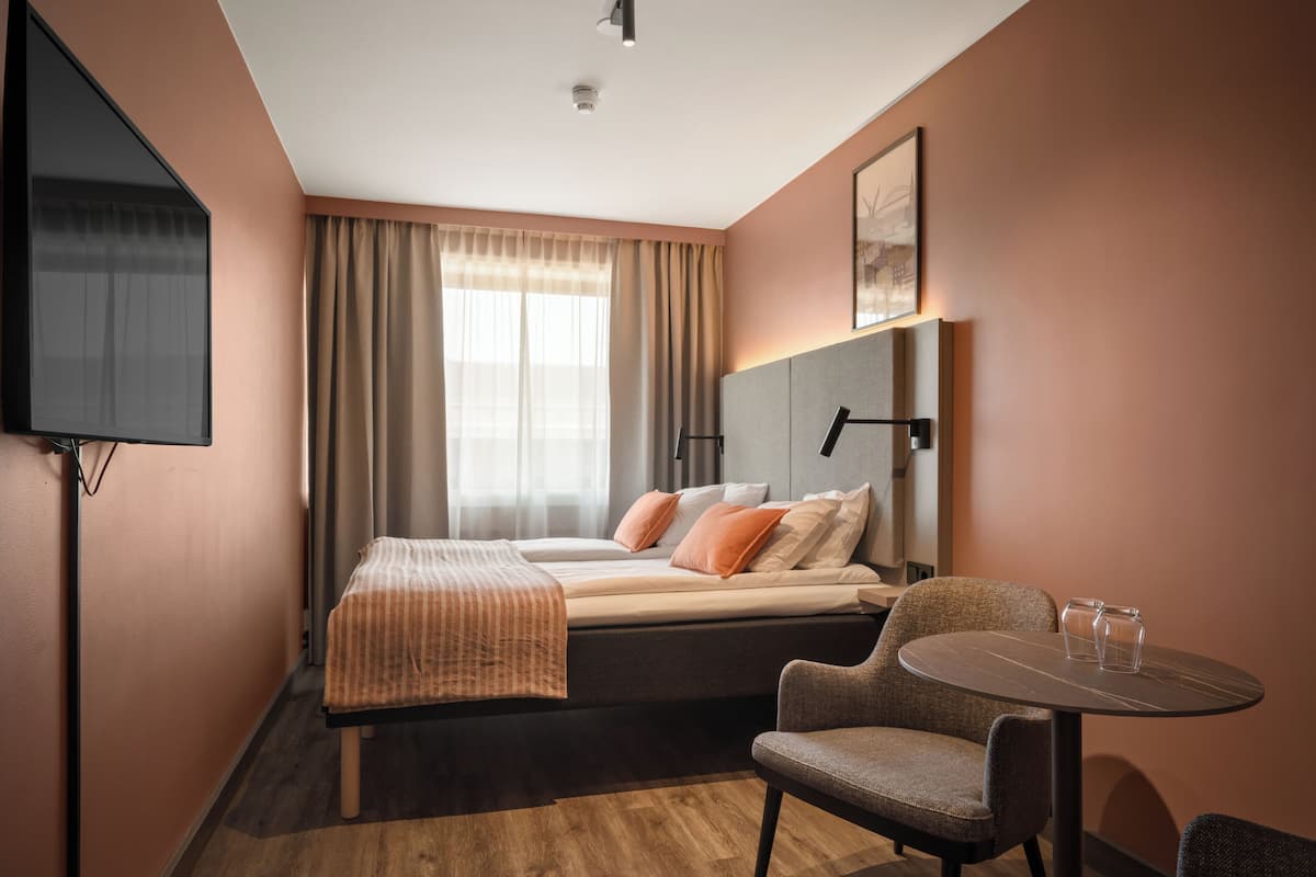 A hotel room with a bed, a chair, a table, a TV, and a window with curtains.