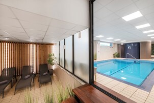 Piscina interior moderna con vistas a la piscina a través de un muro de cristal. Hay sillas de descanso y una mesita con una planta.