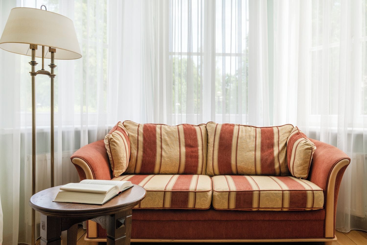 A sofa with striped cushions, a table with a book, and a floor lamp in a room with sheer curtains.