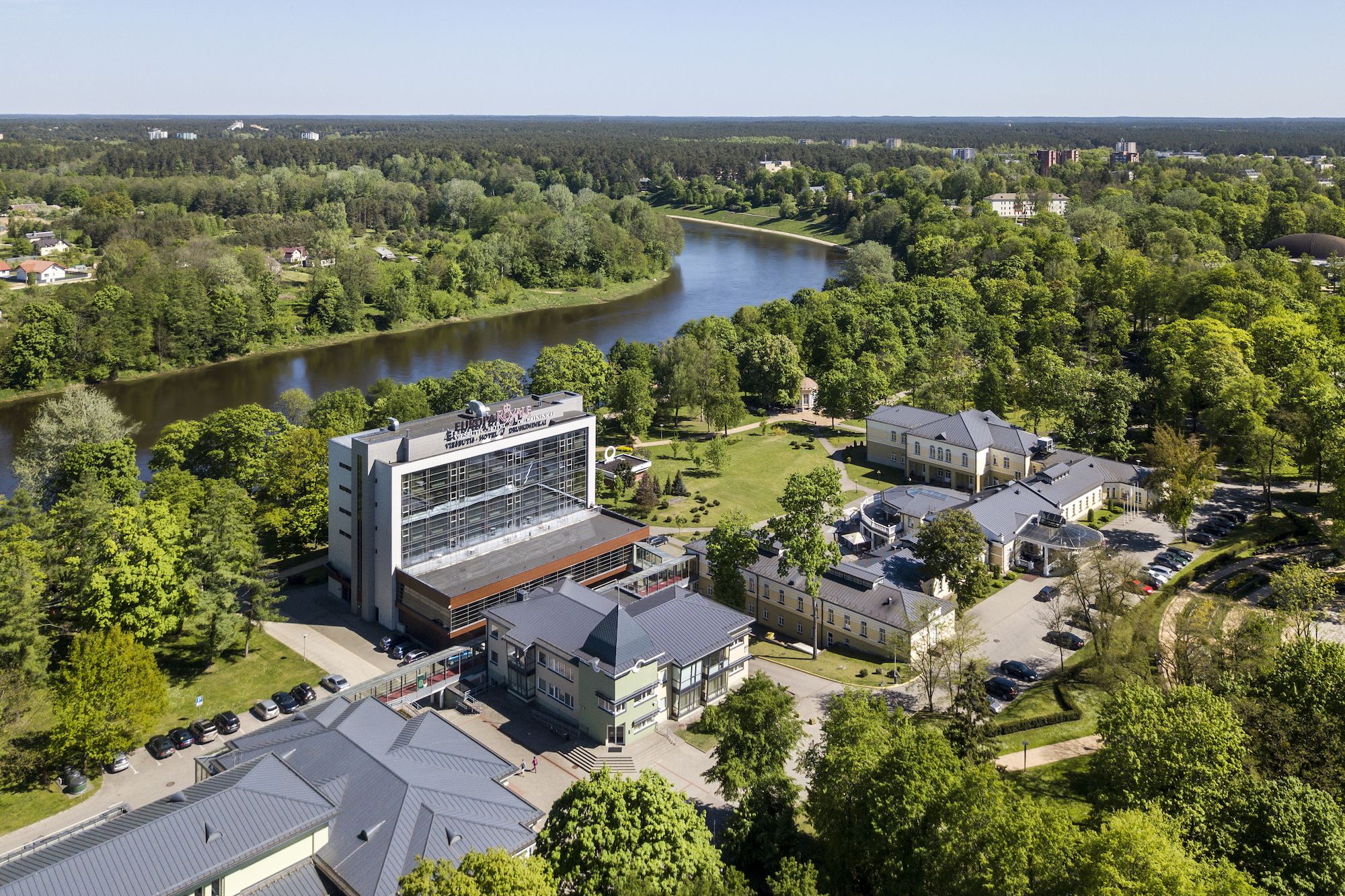 An aerial view of a hotel complex with a modern building, surrounded by lush greenery and a river.