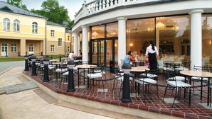 An outdoor cafe with tables and chairs, a white building with large windows, and a brick floor. - Spa Hotel Europa Royale Druskininkai (Druskininkai)