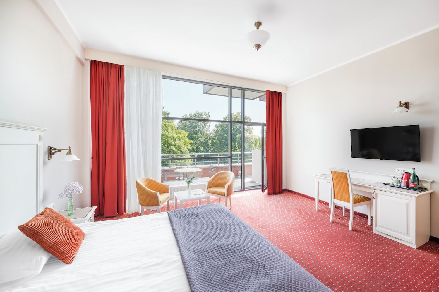A hotel room with a bed, a desk, a chair, a TV, and a balcony with red curtains.