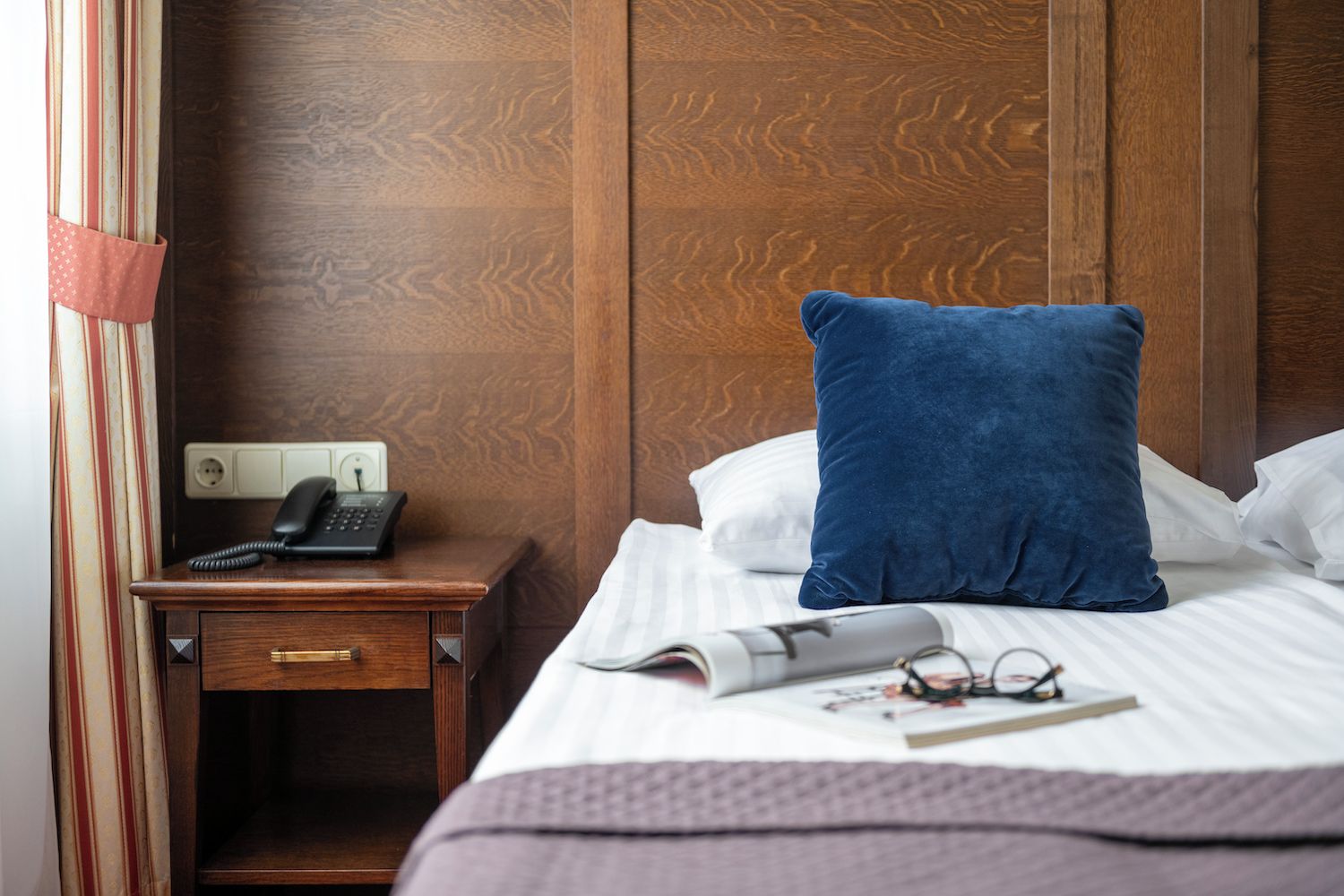 Een hotelkamer met een houten hoofdeinde, een nachtkastje met een telefoon, een blauw kussen, een wit kussen, een opgerold tijdschrift en een paar brillen op het bed.