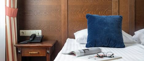 Een hotelkamer met een houten hoofdeinde, een nachtkastje met een telefoon, een blauw kussen, een wit kussen, een opgerold tijdschrift en een paar brillen op het bed.