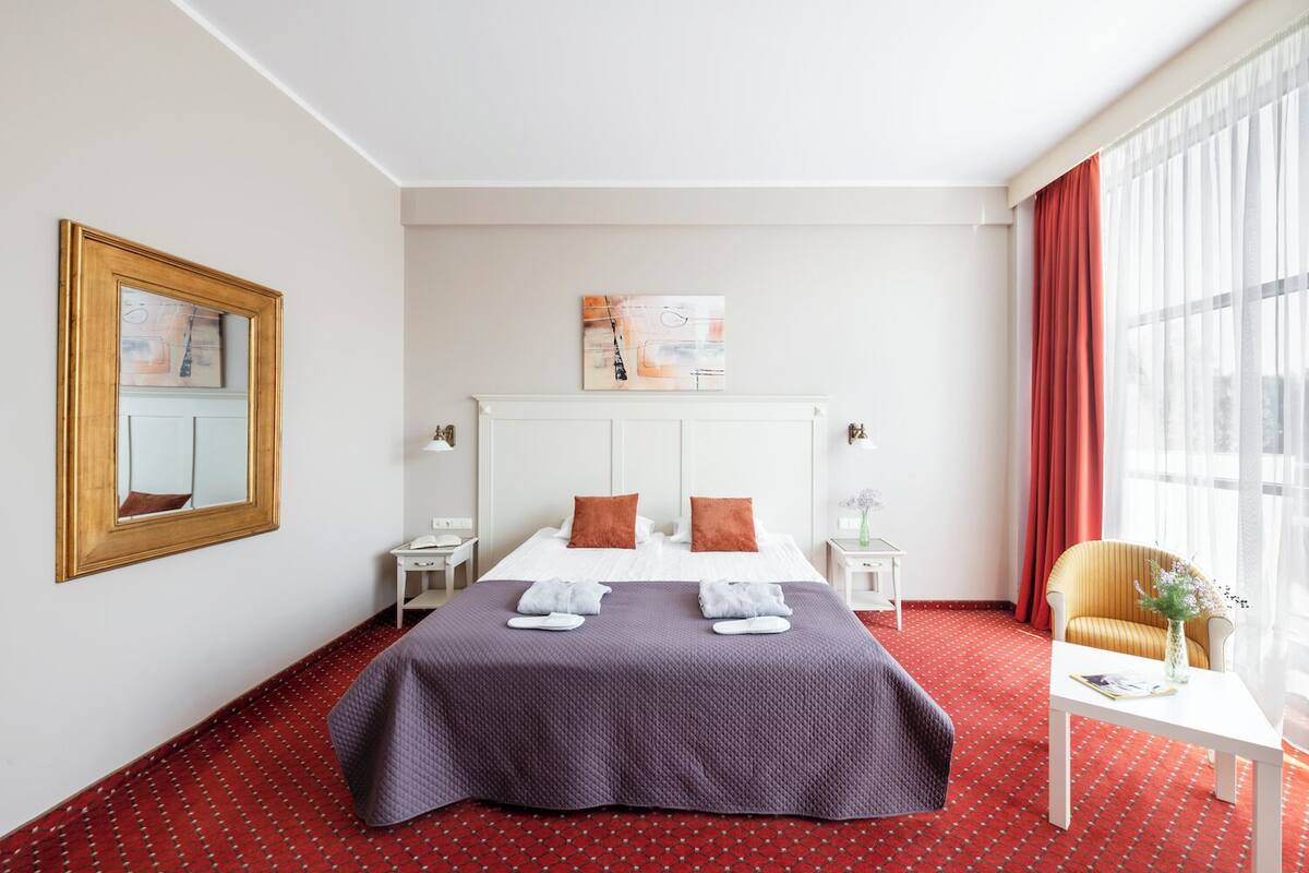 Une chambre d’hôtel avec un grand lit, une table de chevet, une chaise, une fenêtre avec des rideaux et un tableau au mur.