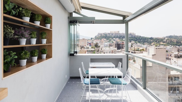 A balcony with a table and chairs, wooden shelves with potted plants, and a view of a cityscape with a hill in the background. - Evripides Hotel (Athens)