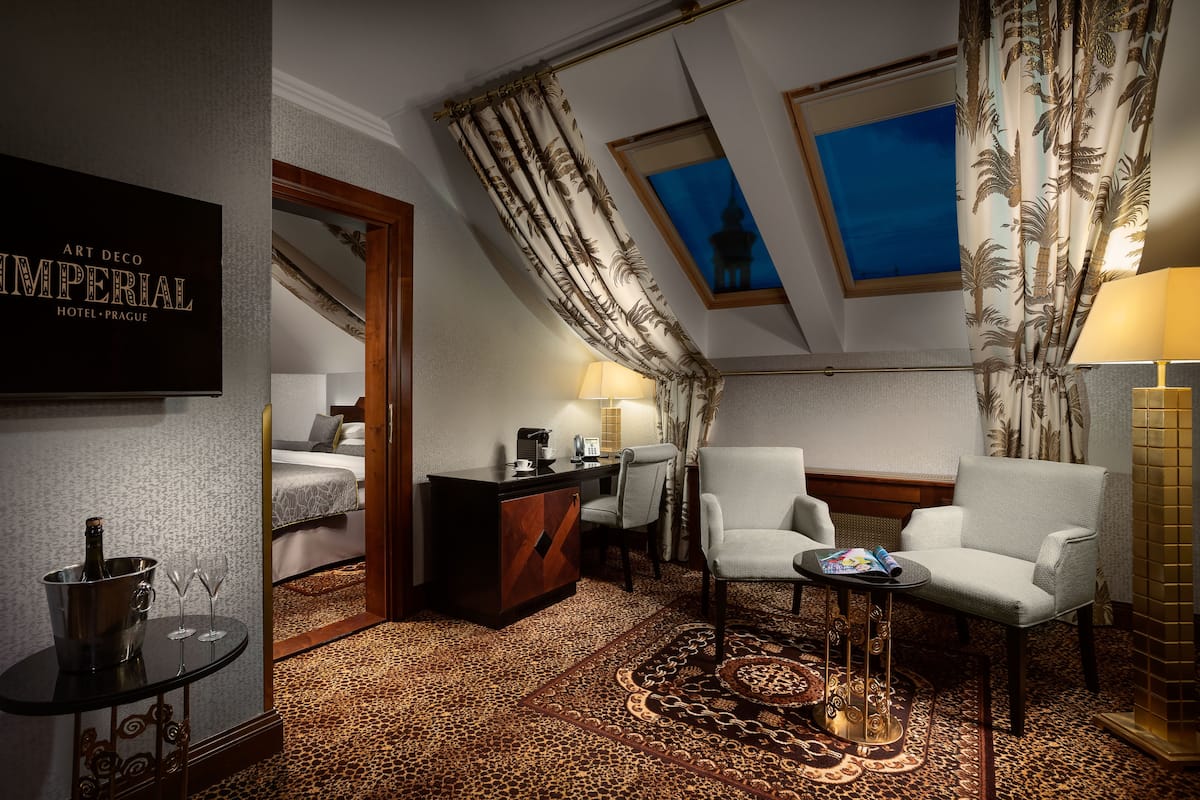 Hotelkamer met een bed, een bureau, twee fauteuils, een kleine tafel met lamp, een minibar en uitzicht op een gebouw door een raam.