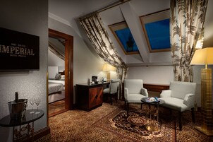 Habitación de hotel con cama, escritorio, dos sillones, una mesita de noche con lámpara, un minibar y vistas a un edificio a través del ventanal.