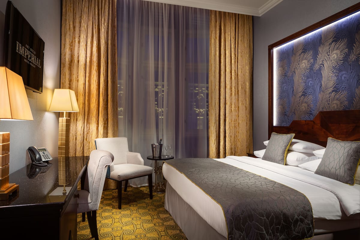 Hotelkamer met een bed, een bureau met telefoon, een stoel, een tafeltje met een glas en uitzicht op de stad door gordijnen.