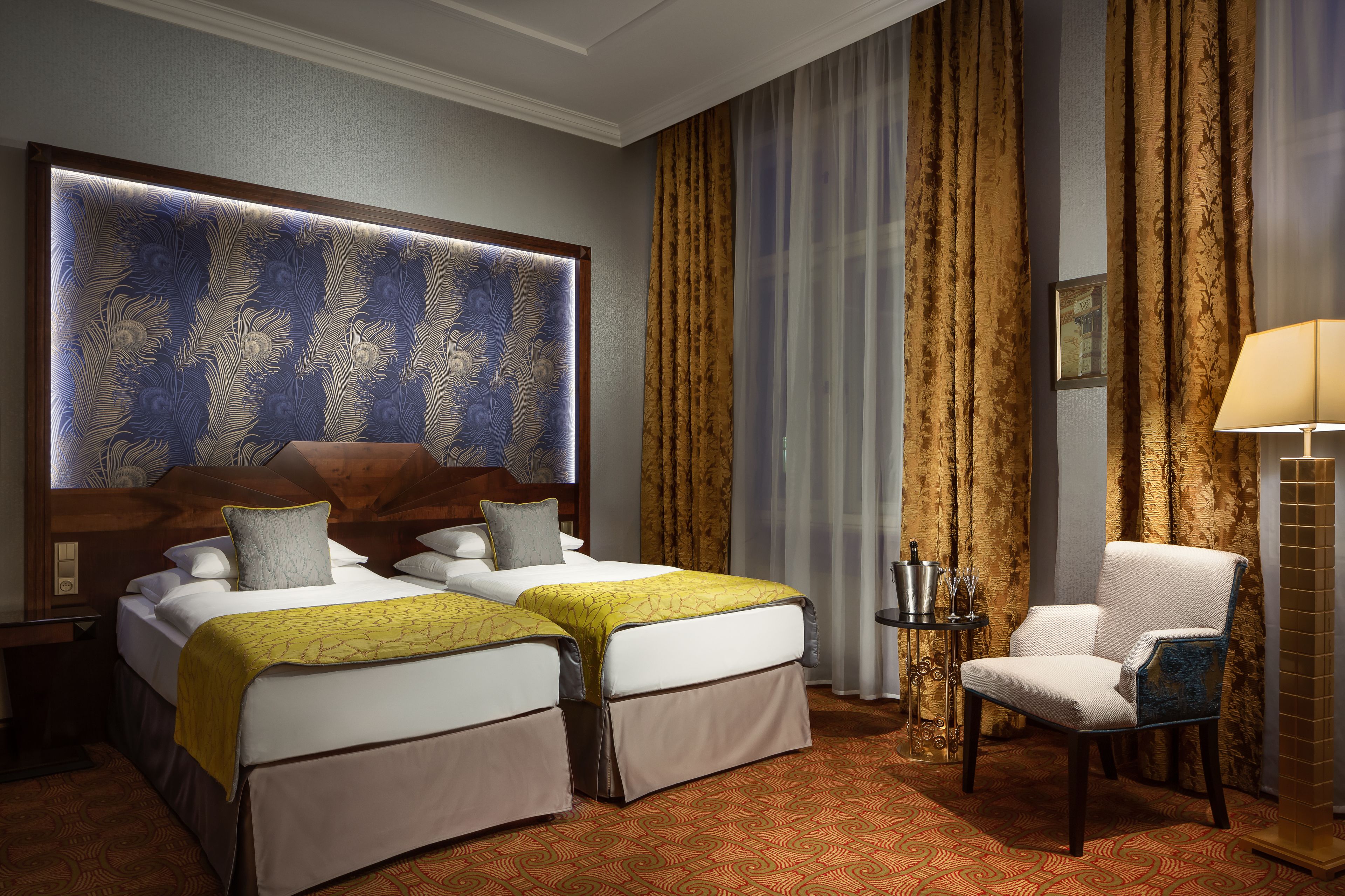 Una camera d'albergo con due letti, una grande testiera decorativa, una sedia, un comodino e una lampada.