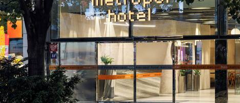 Entrada de hotel com fachada de vidro, uma placa digital exibindo o nome do hotel e uma área de recepção visível pelo vidro.