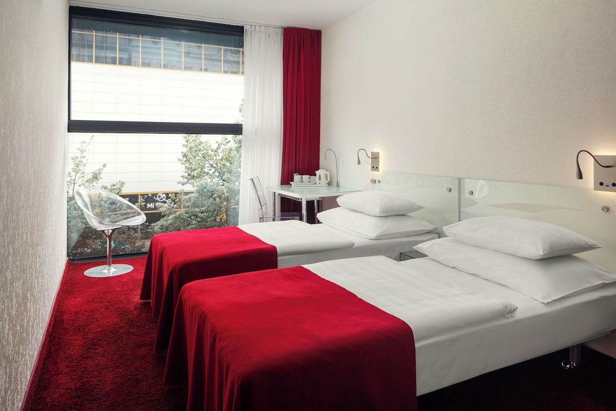 Quarto de hotel com duas camas, um tapete vermelho, uma mesa de vidro com chaleira e vista para áreas verdes.