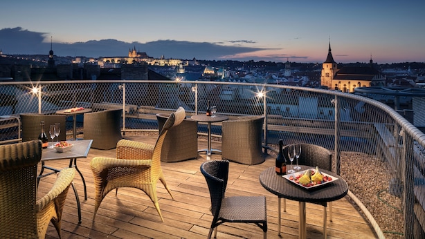 Une terrasse sur le toit, aménagée avec des meubles en osier, des tables et des chaises, d’où l’on peut admirer un panorama sur la ville, avec ses bâtiments historiques et le clocher d’une église.