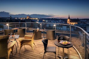 A rooftop terrace with wicker furniture, tables, and chairs, overlooking a cityscape with historic buildings and a church spire. - Metropol Design Hotel Old Town (Prague)