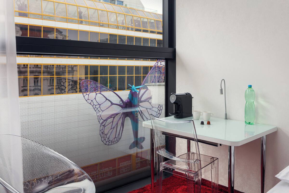 Quarto moderno com mesa de vidro, cadeiras transparentes, tapete vermelho, máquina de café e uma garrafa de água.