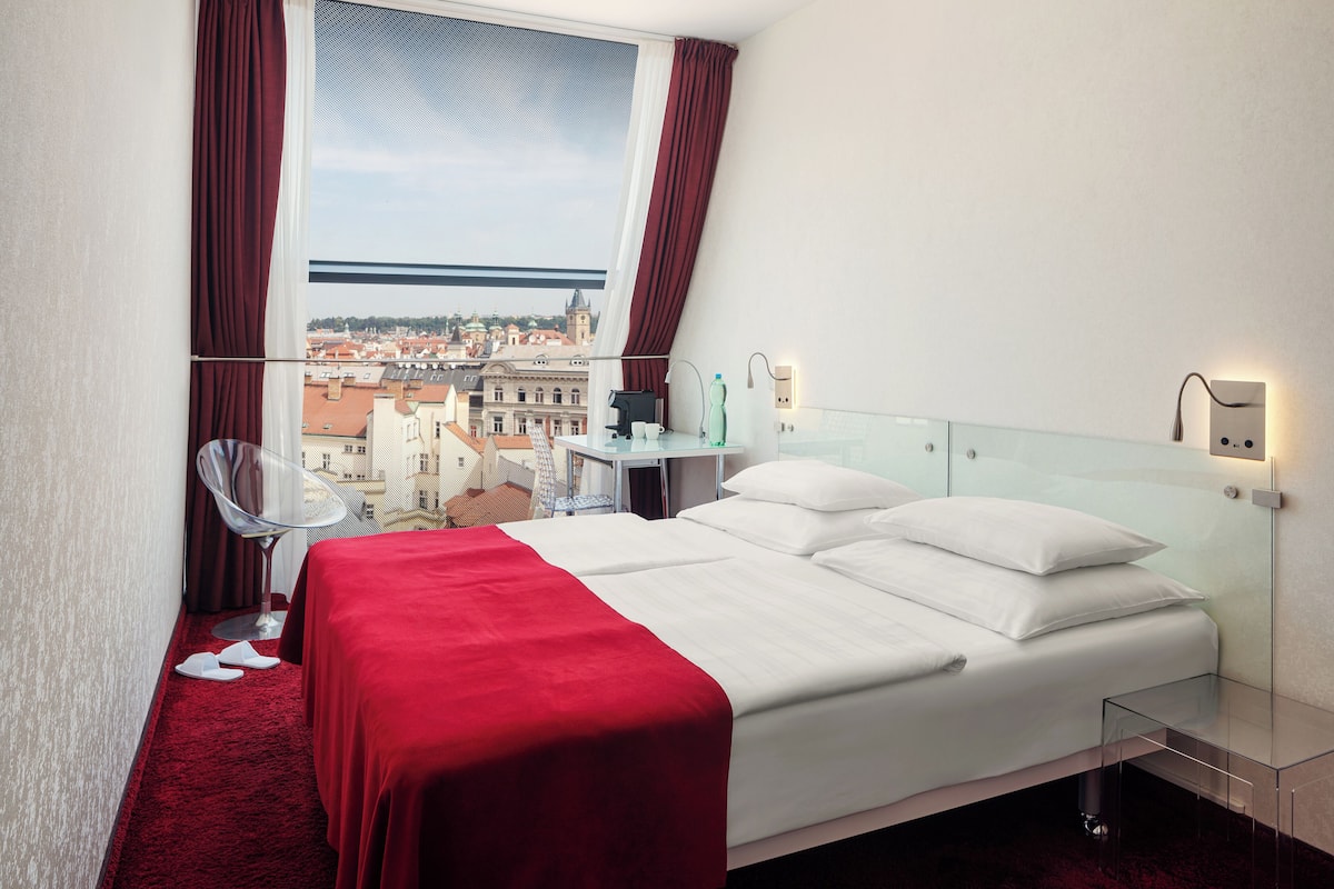 Quarto de hotel com uma cama grande, mesa de cabeceira de vidro, uma cadeira e vista para a cidade.