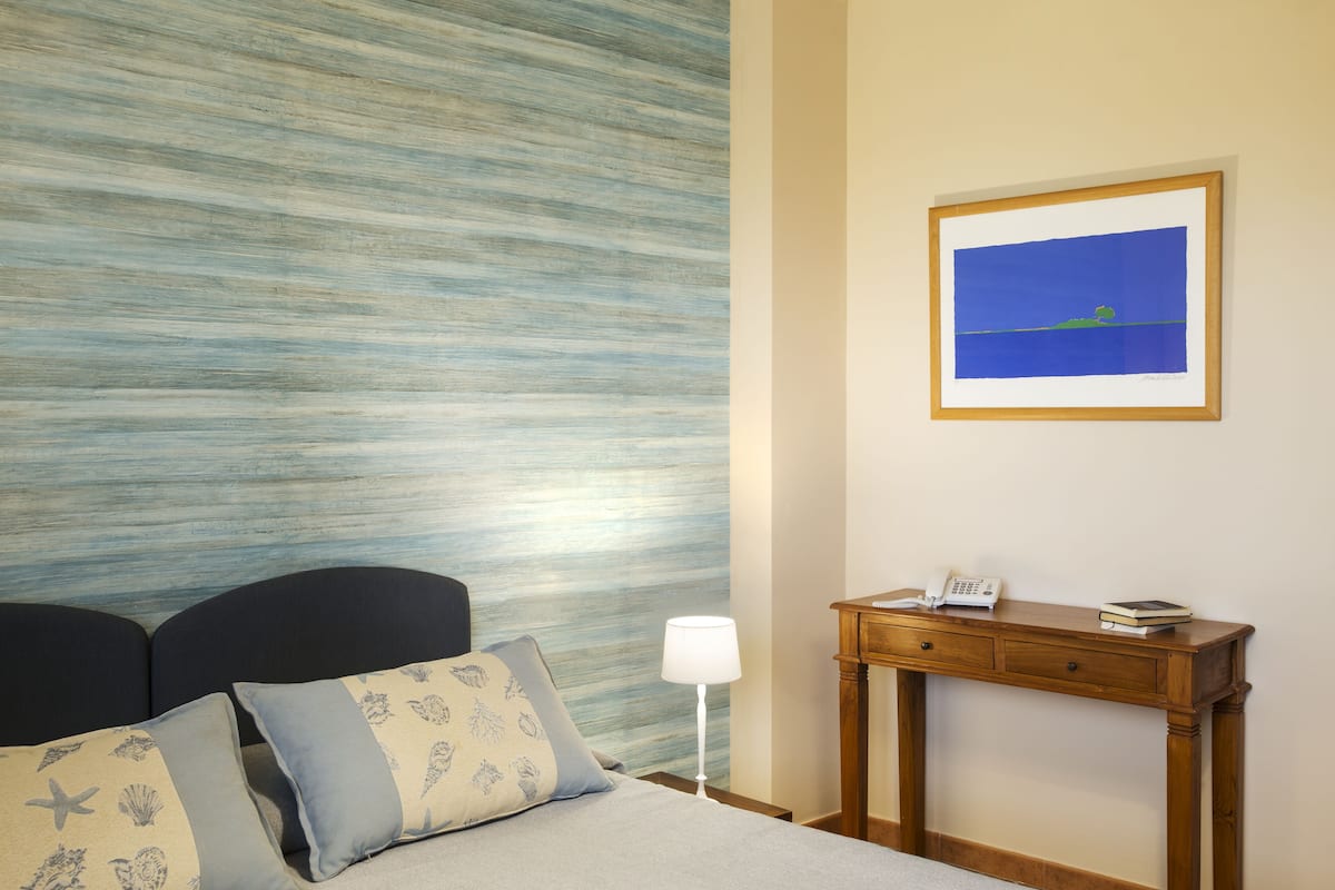 Habitación de hotel con cama, mesita de noche, lámpara, escritorio de madera y un cuadro enmarcado en la pared.