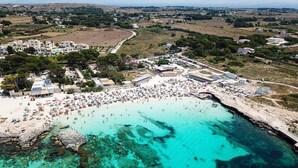 An aerial view of a coastal area with a beach, buildings, and clear turquoise waters. - FAVIGNANA HOTEL Concept Holiday (Favignana)
