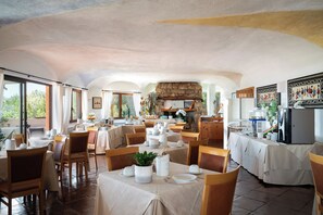 A restaurant with multiple tables set for dining, a stone fireplace, and a view of the outdoors. - Hotel Valdiola (Arzachena)