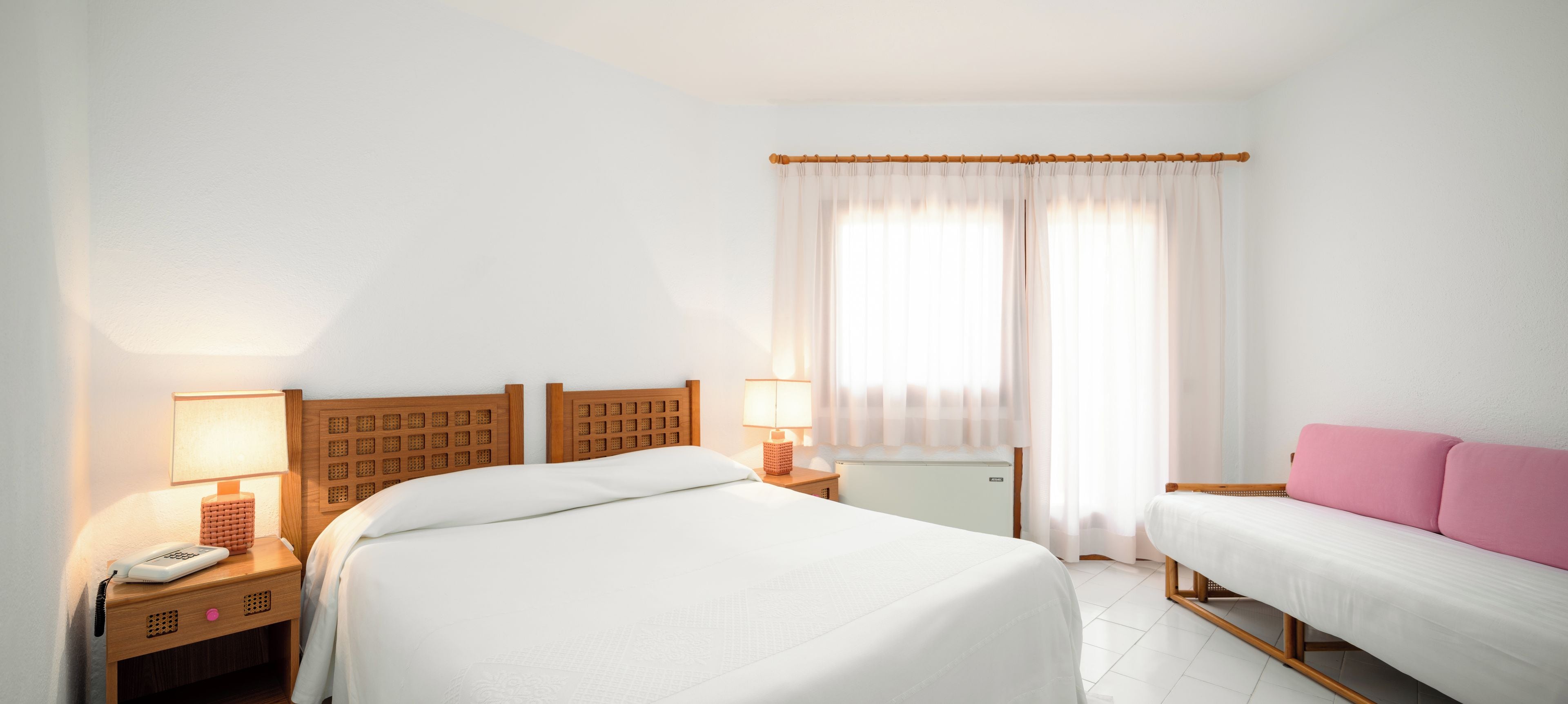 A hotel room with two beds, a bedside table with a lamp, a telephone, and a window with curtains.