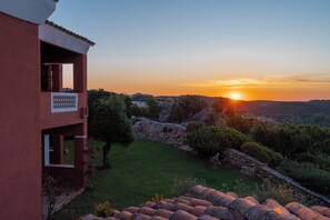 A red building with a balcony overlooking a grassy area and a sunset. - Hotel Valdiola (Arzachena)