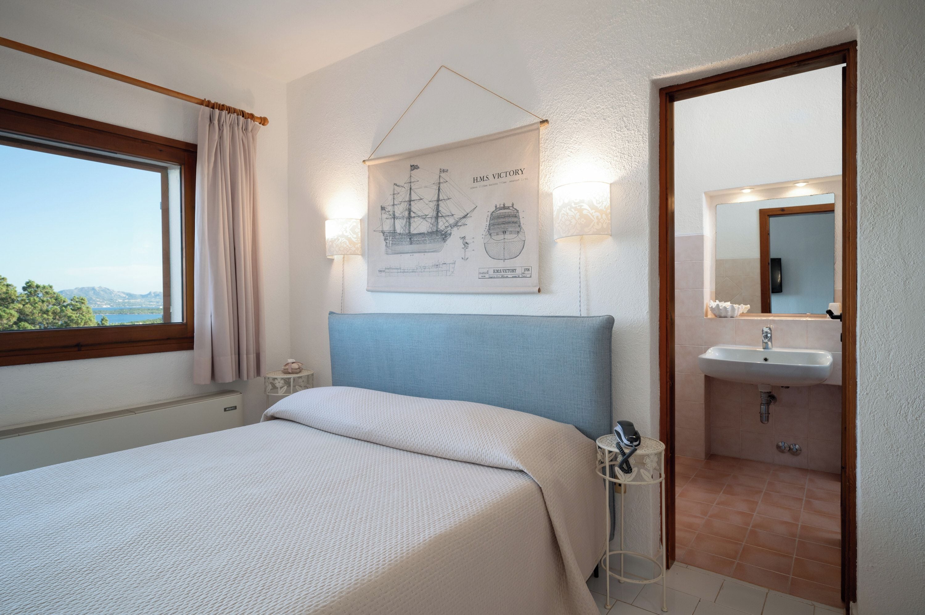 A bedroom with a bed, a window with curtains, a wall-mounted lamp, a framed ship illustration, and a bathroom with a sink and mirror.