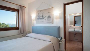 A bedroom with a bed, a window with curtains, a wall-mounted lamp, a framed ship illustration, and a bathroom with a sink and mirror.