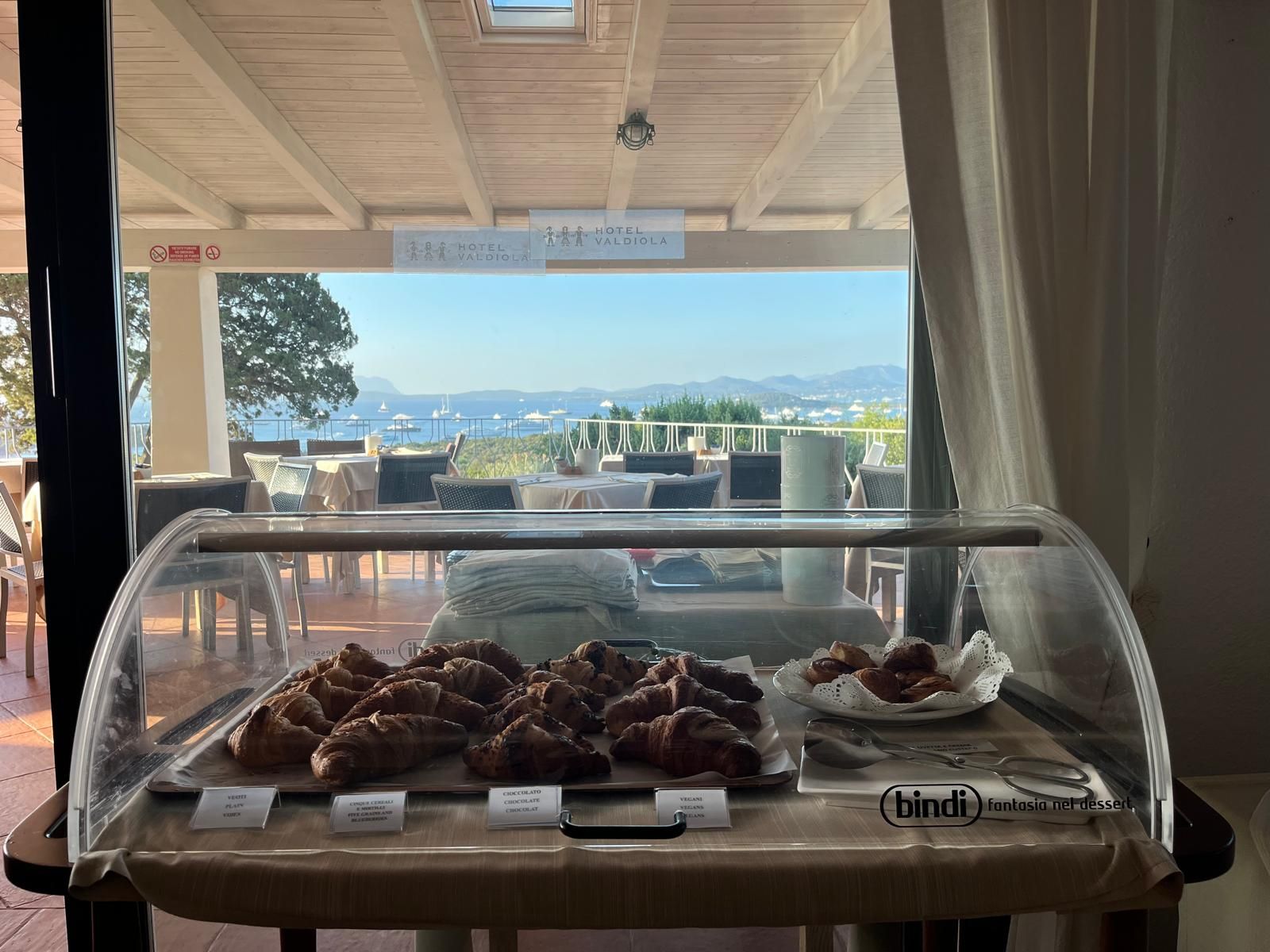 Mostrador de panadería con croissants y opciones veganas, con vista a una terraza con mesas al aire libre y una vista panorámica.