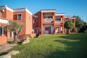 A row of terracotta-colored buildings with balconies, a grassy lawn, and a clear blue sky. - Hotel Valdiola (Arzachena)