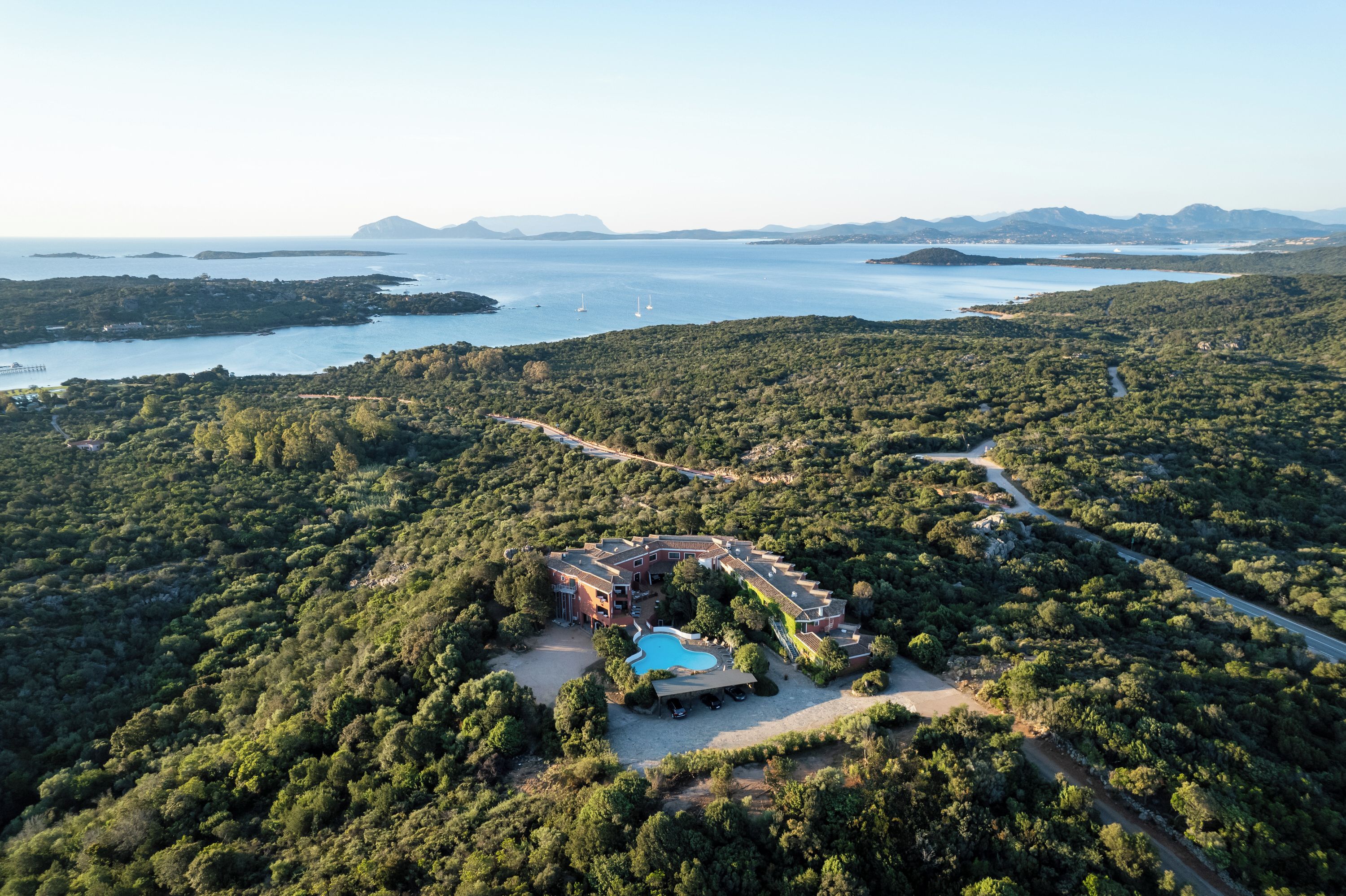An aerial view of a resort with a pool, surrounded by dense forest and a clear view of the sea.