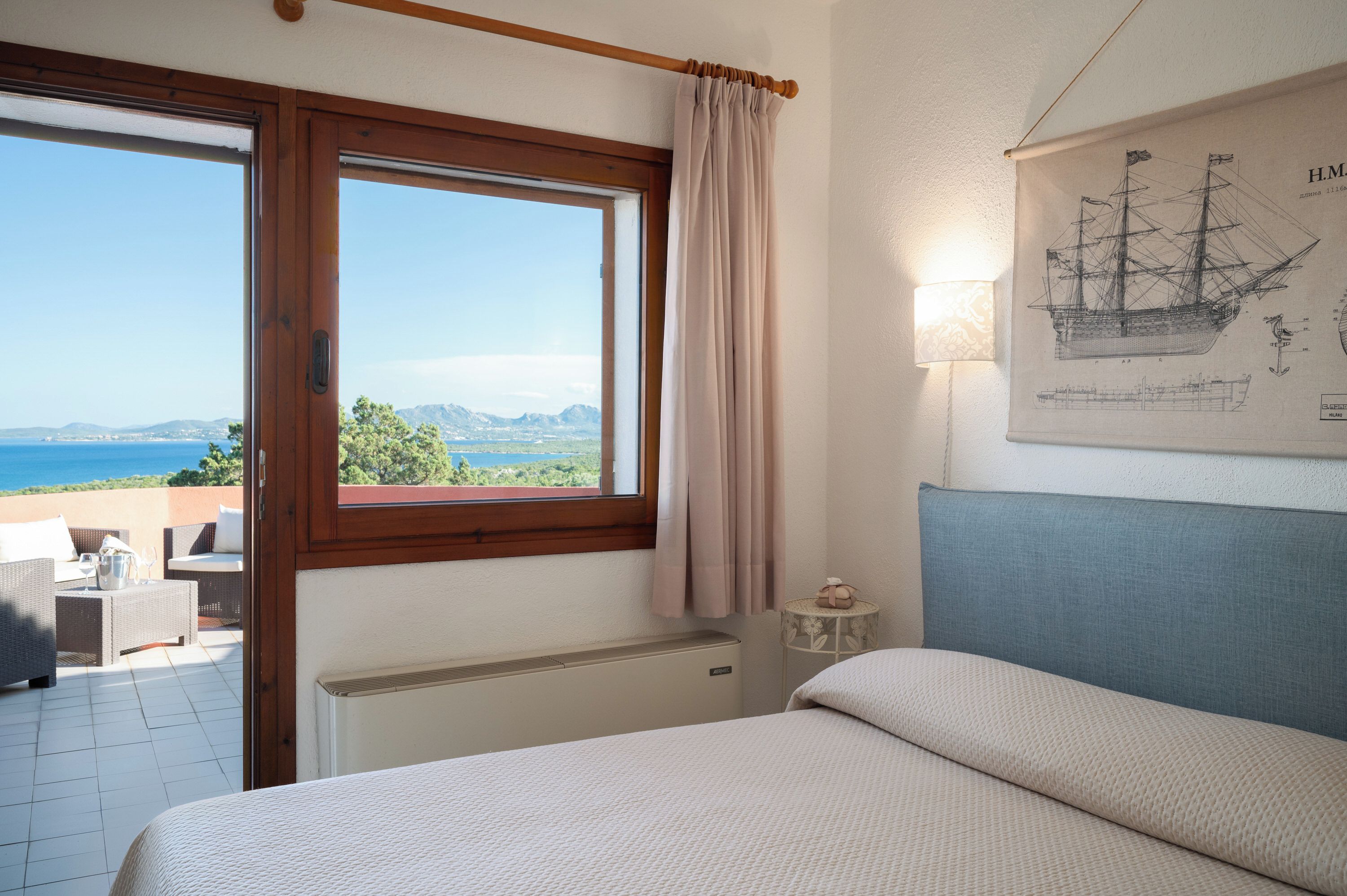 A bedroom with a bed, a window with curtains, a radiator, a wall-mounted lamp, and a framed map.