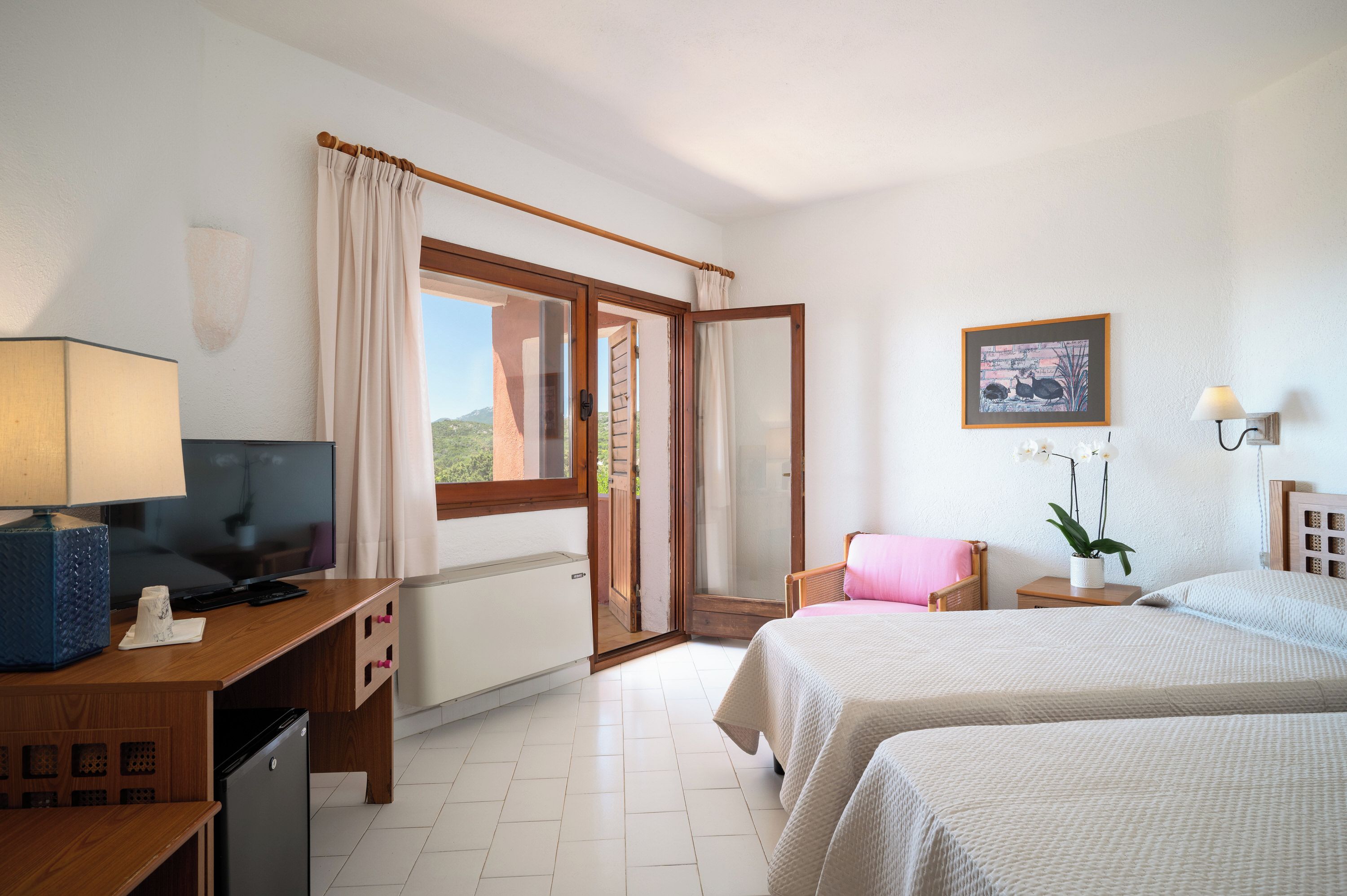 Habitación de hotel con cama, escritorio con televisor, una silla, ventana con cortinas e imagen colgada en la pared.
