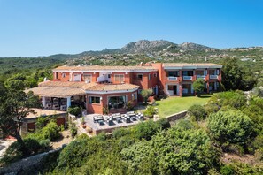A large, multi-story building with a red facade, a terrace with outdoor furniture, and a view of a mountainous landscape in the background. - Hotel Valdiola (Arzachena)