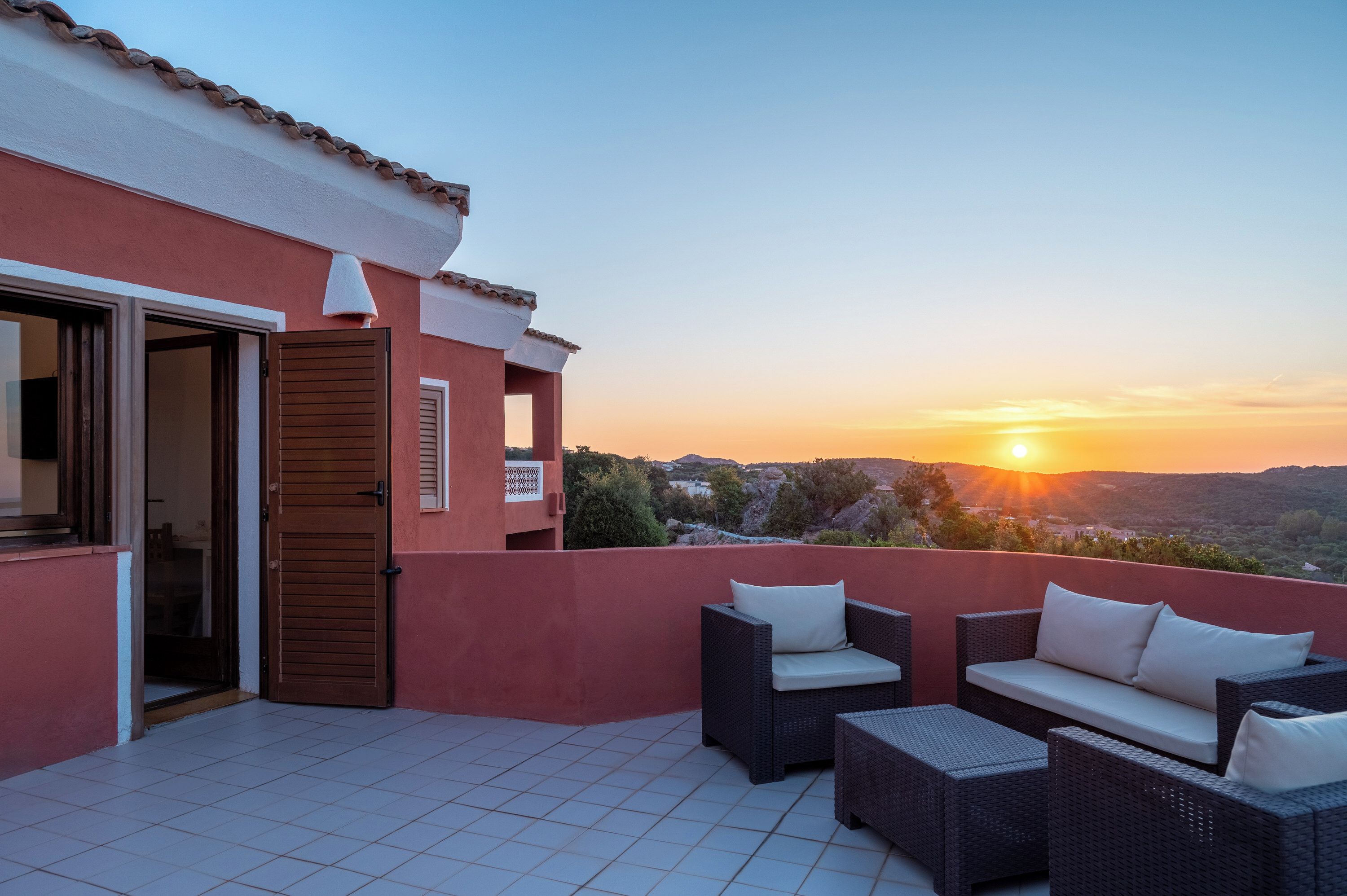 A terrace with wicker furniture and a view of the sunset over a mountainous landscape.