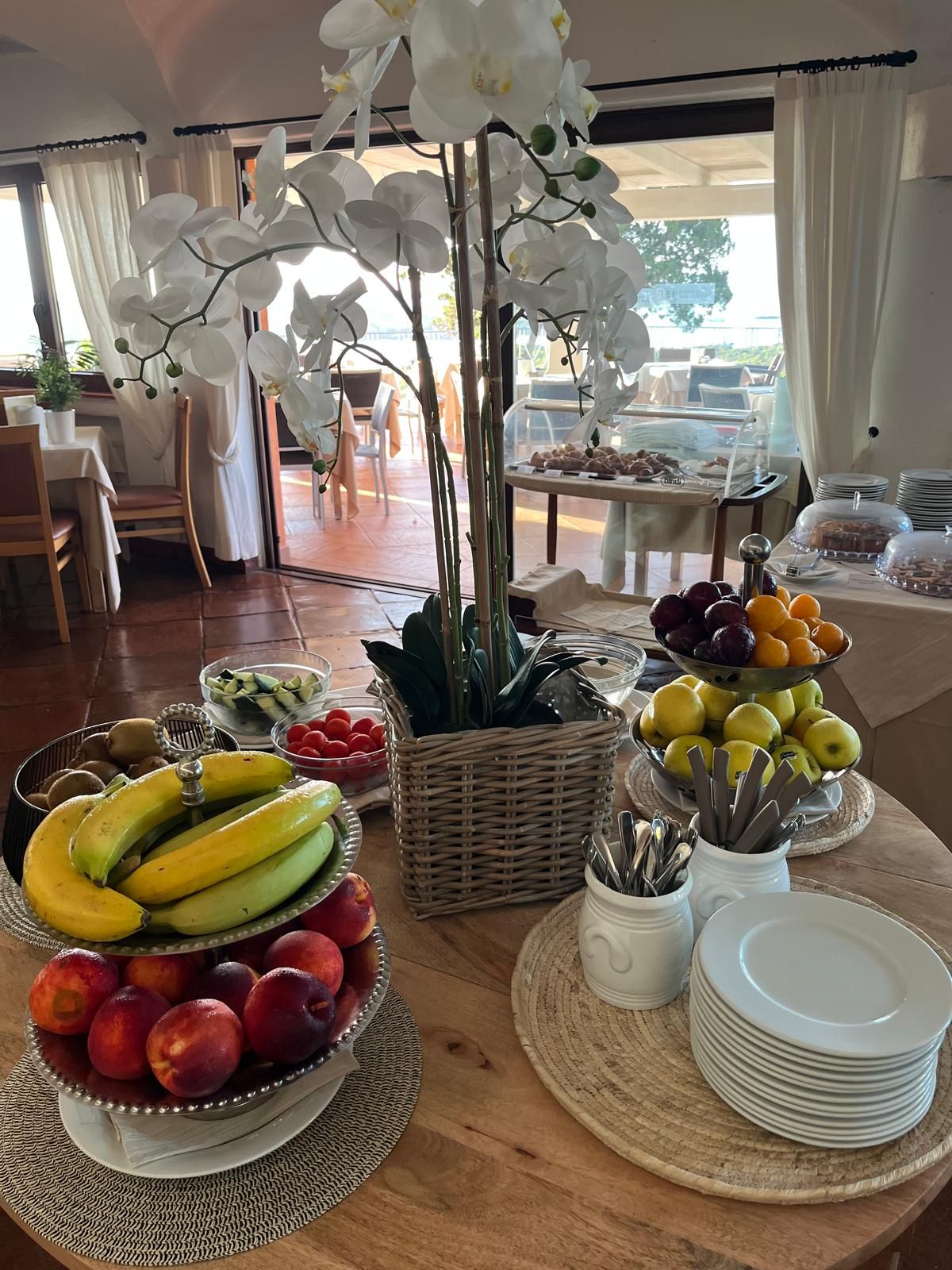 A breakfast buffet with fruits, a wicker basket, and a table with plates and utensils.