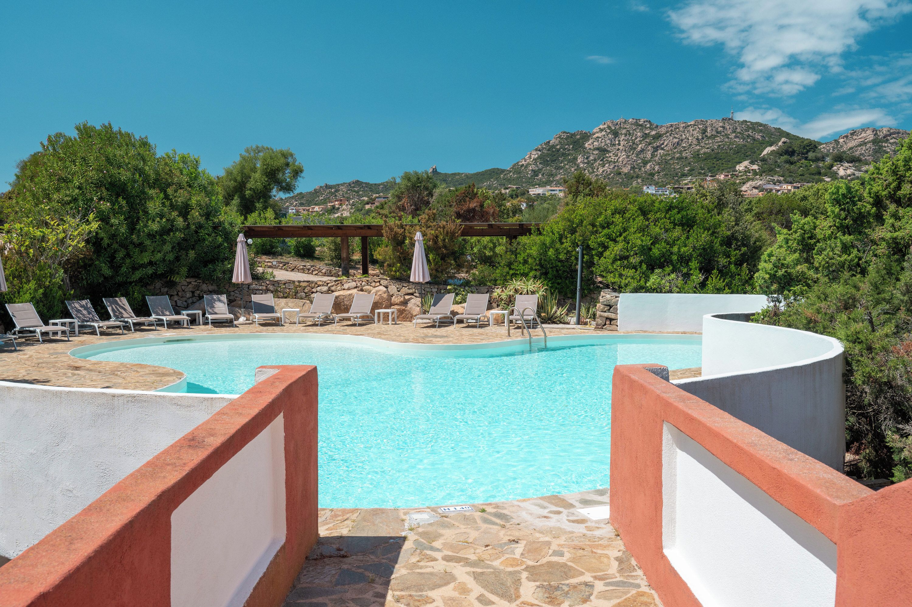 Una piscina con lettini e ombrelloni, immersa nel verde e con vista sulle montagne.