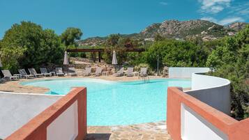Una piscina con lettini e ombrelloni, immersa nel verde e con vista sulle montagne.