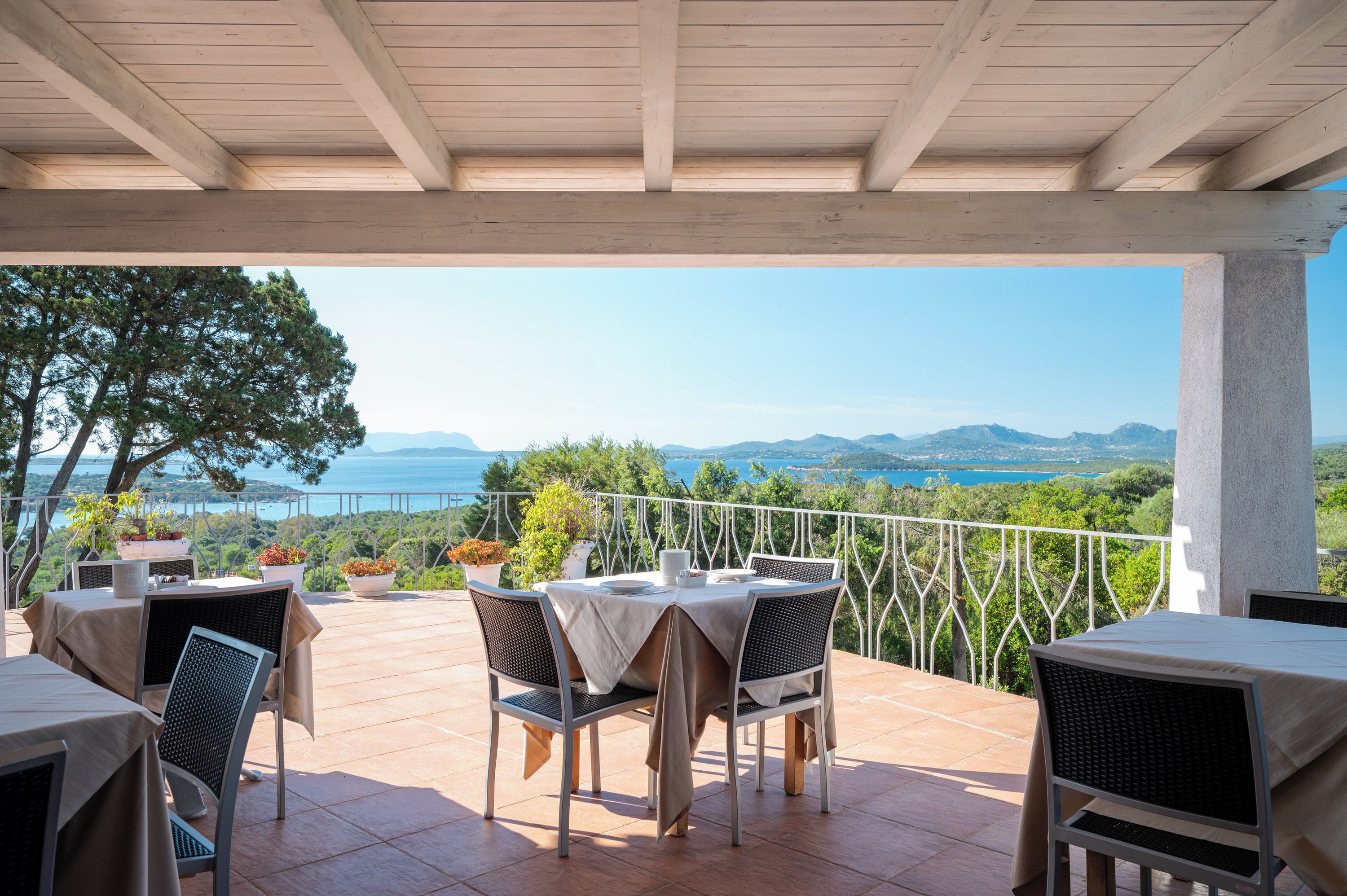 Une terrasse extérieure avec des tables et des chaises, offrant une vue panoramique sur un paysage magnifique, avec des montagnes et une étendue d’eau.
