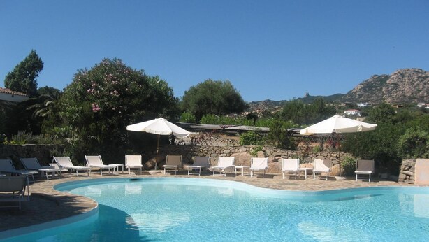 A clear blue swimming pool with white lounge chairs and umbrellas, surrounded by greenery and a stone wall. - Hotel Valdiola (Arzachena)