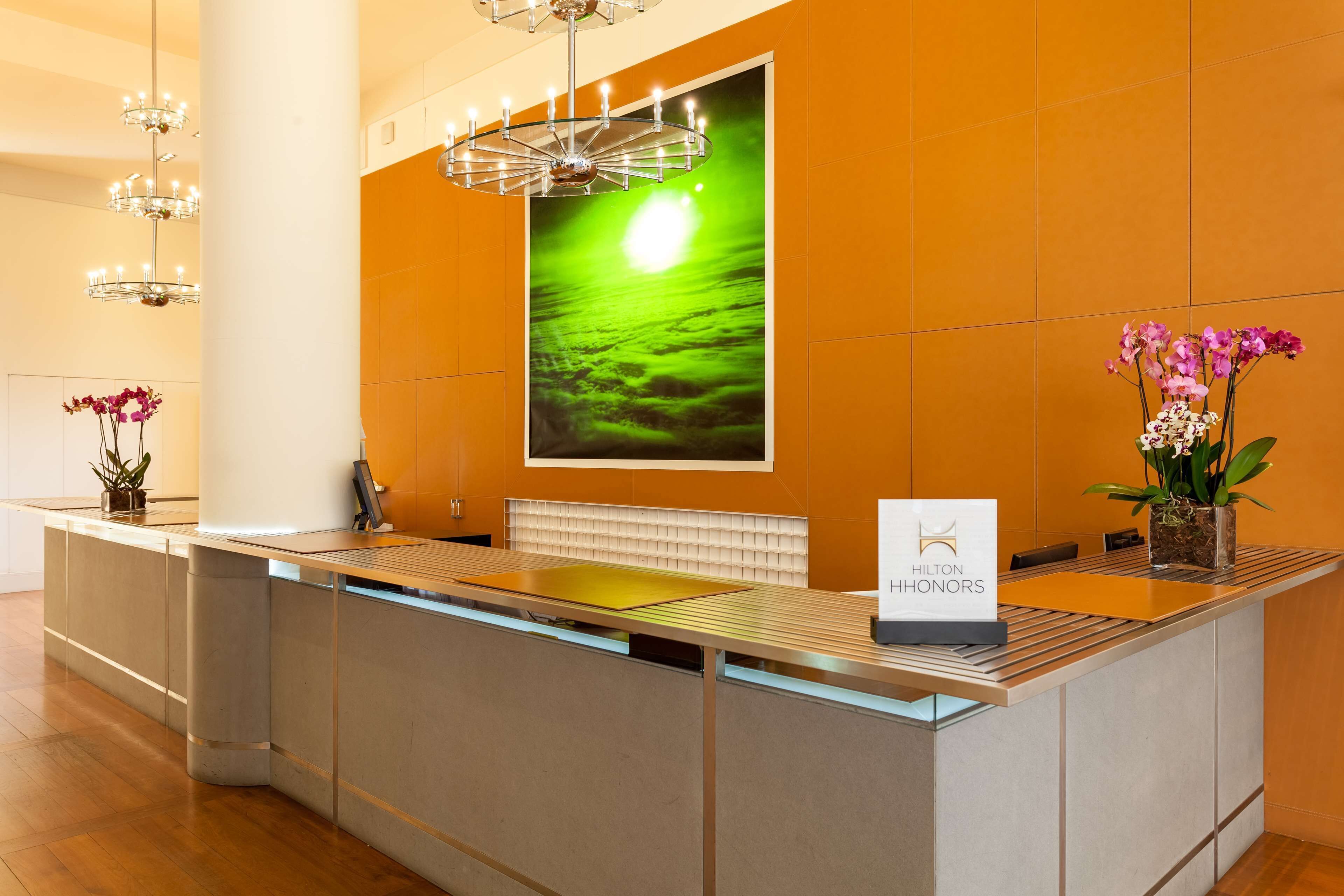 Un mostrador de recepción de hotel con diseño moderno, un amplio ventanal con vistas a un paisaje verde, y un cartel que indica 'Hilton Honors'.