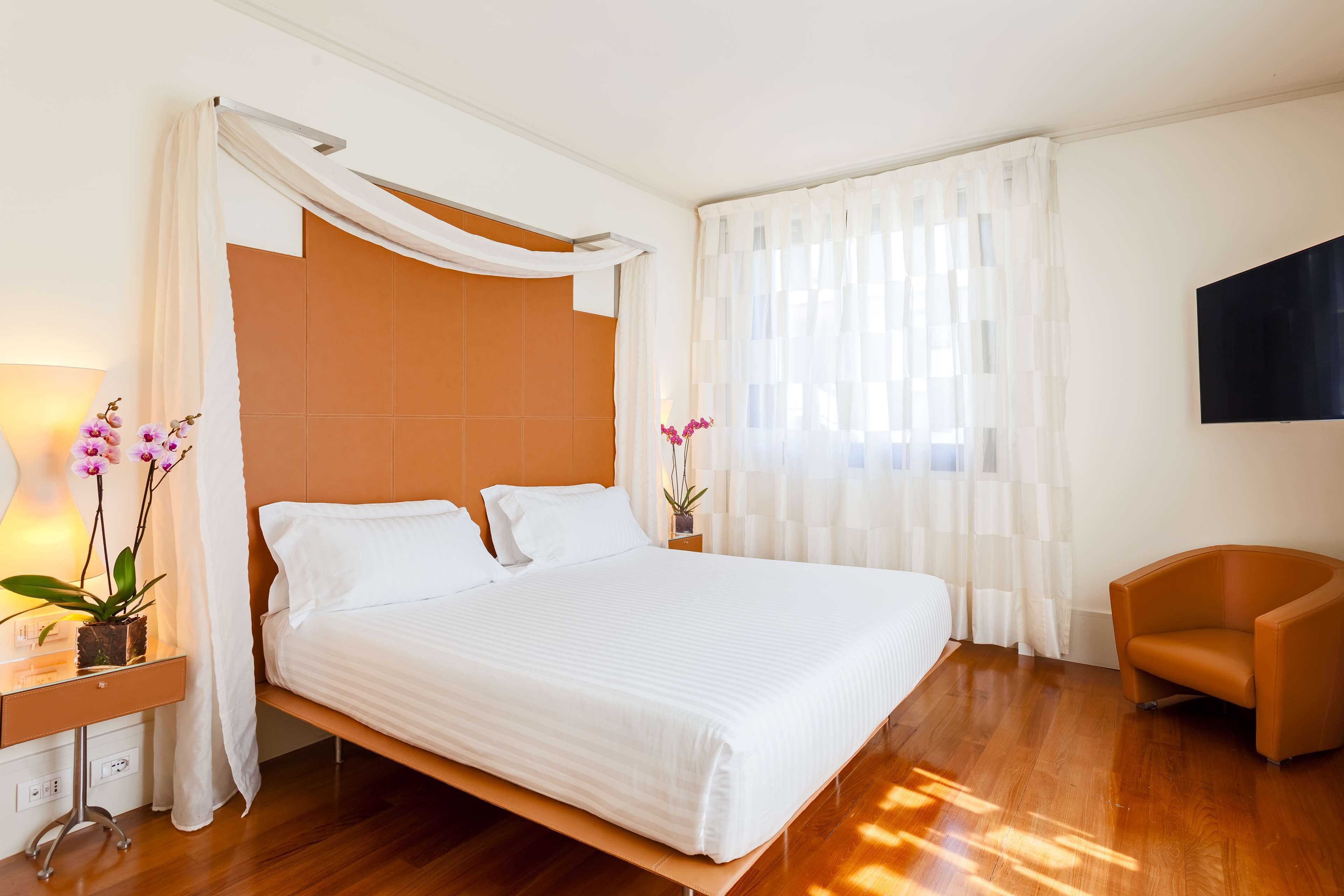 A hotel room with a large bed, a bedside table with a lamp and flowers, a chair, and a TV on the wall.