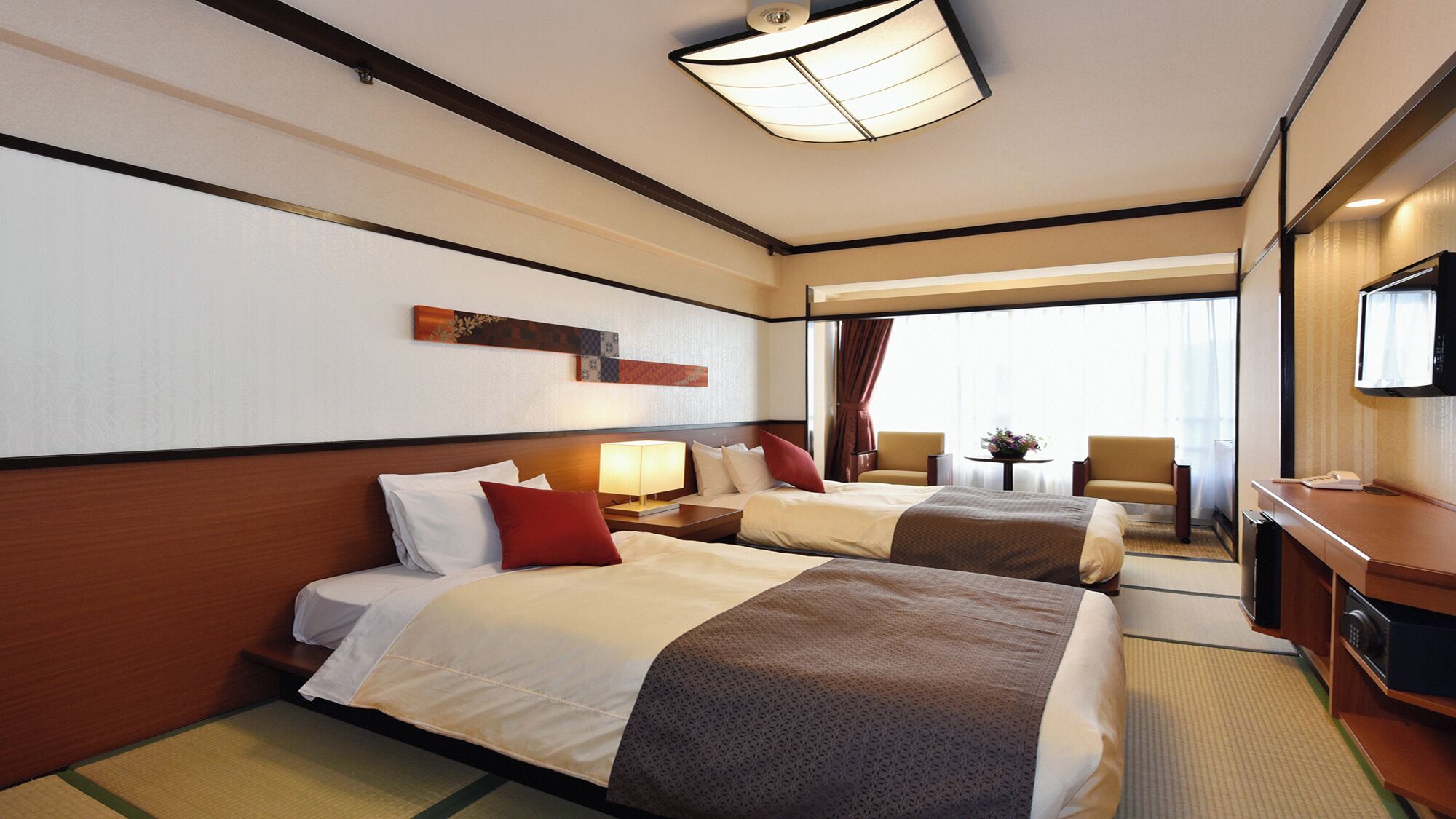 A hotel room with two beds, a desk with a television, a lamp, and a view of the outside.