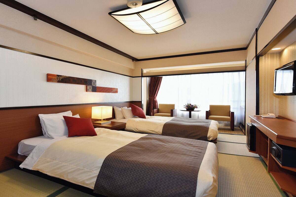 A hotel room with two beds, a desk with a television, a lamp, and a view of the outside.