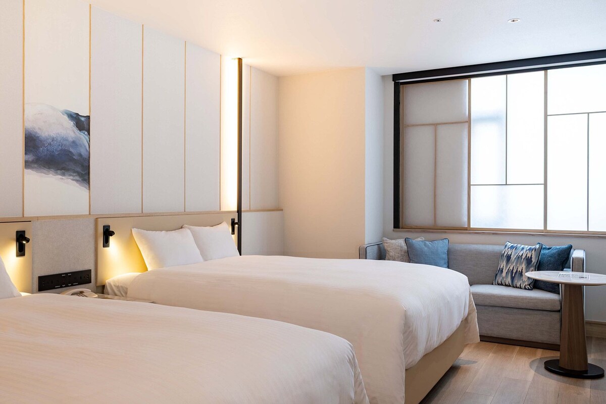 A hotel room with two beds, a sofa, a small table, and a large window.