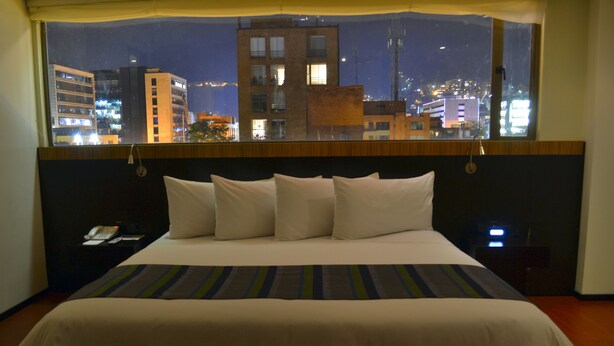 A hotel room with a large bed, a wooden headboard, and a view of the city at night. - Hotel Madisson Inn Luxury By Geh Suites (Bogotá)