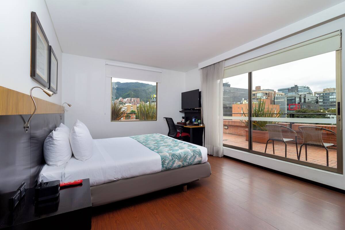 A modern hotel room with a bed, a desk, a TV, and a balcony with chairs.