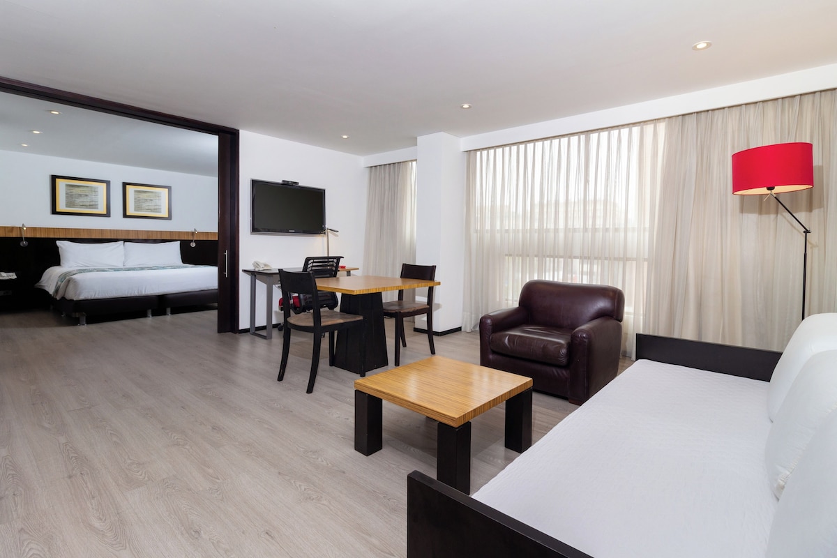 A hotel room with a bed, a desk, a chair, a sofa, a TV, and a lamp.
