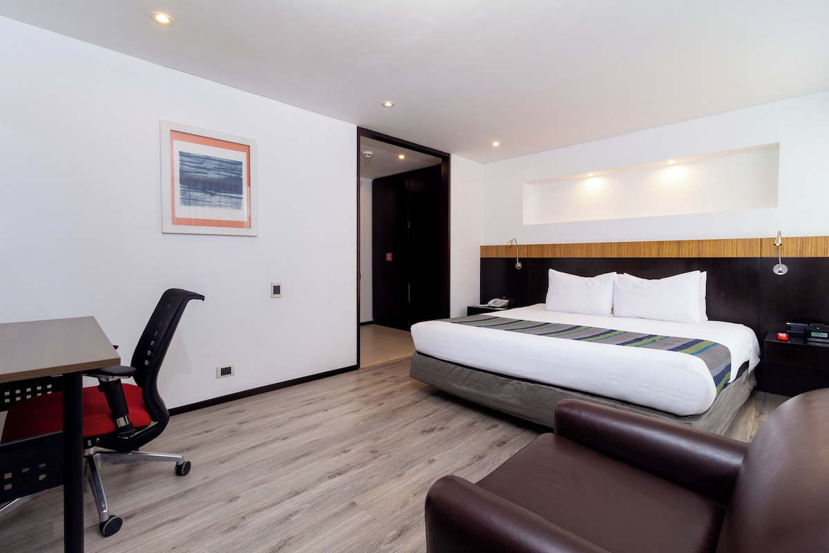 A hotel room with a bed, a desk, a chair, and a bedside table.