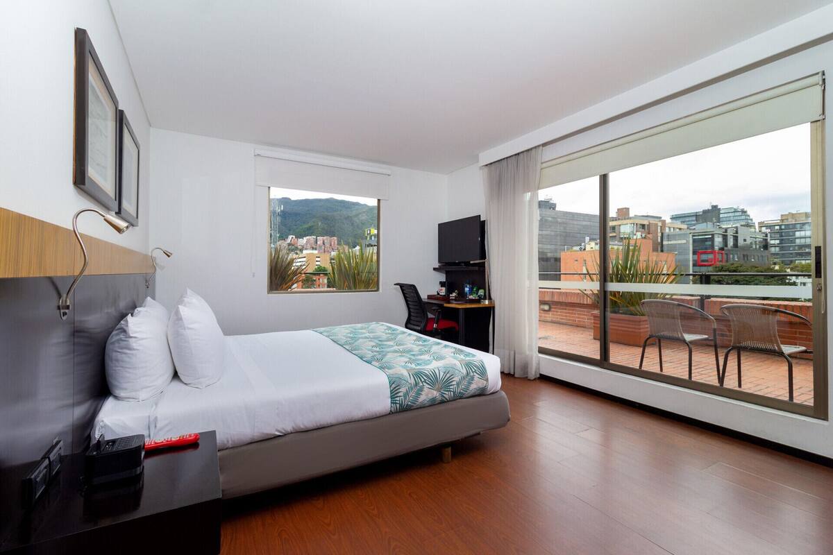 A modern hotel room with a bed, a desk, a TV, and a balcony with chairs.
