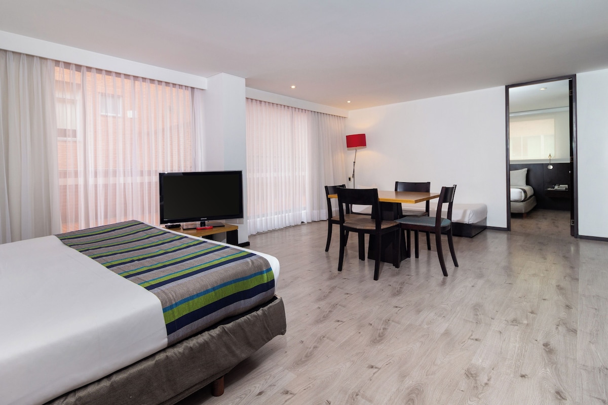 A hotel room with a bed, a TV, a desk with chairs, and a view of the outside through large windows.